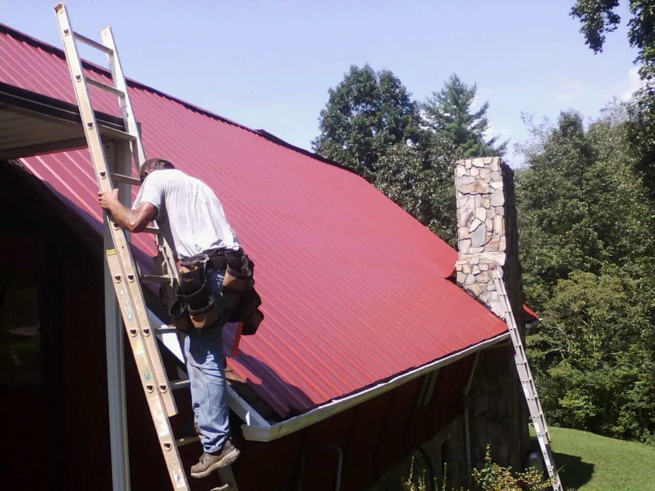Roofer on a ladder beside a red metal roof and stone chimney — Soffit & Fascia Repair by Roofing Brothers in Lakewood